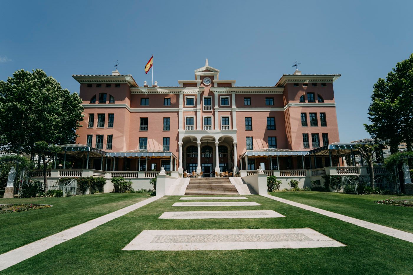 A grand pink historic mansion with ornate architecture and symmetrical design sits atop manicured lawn steps.