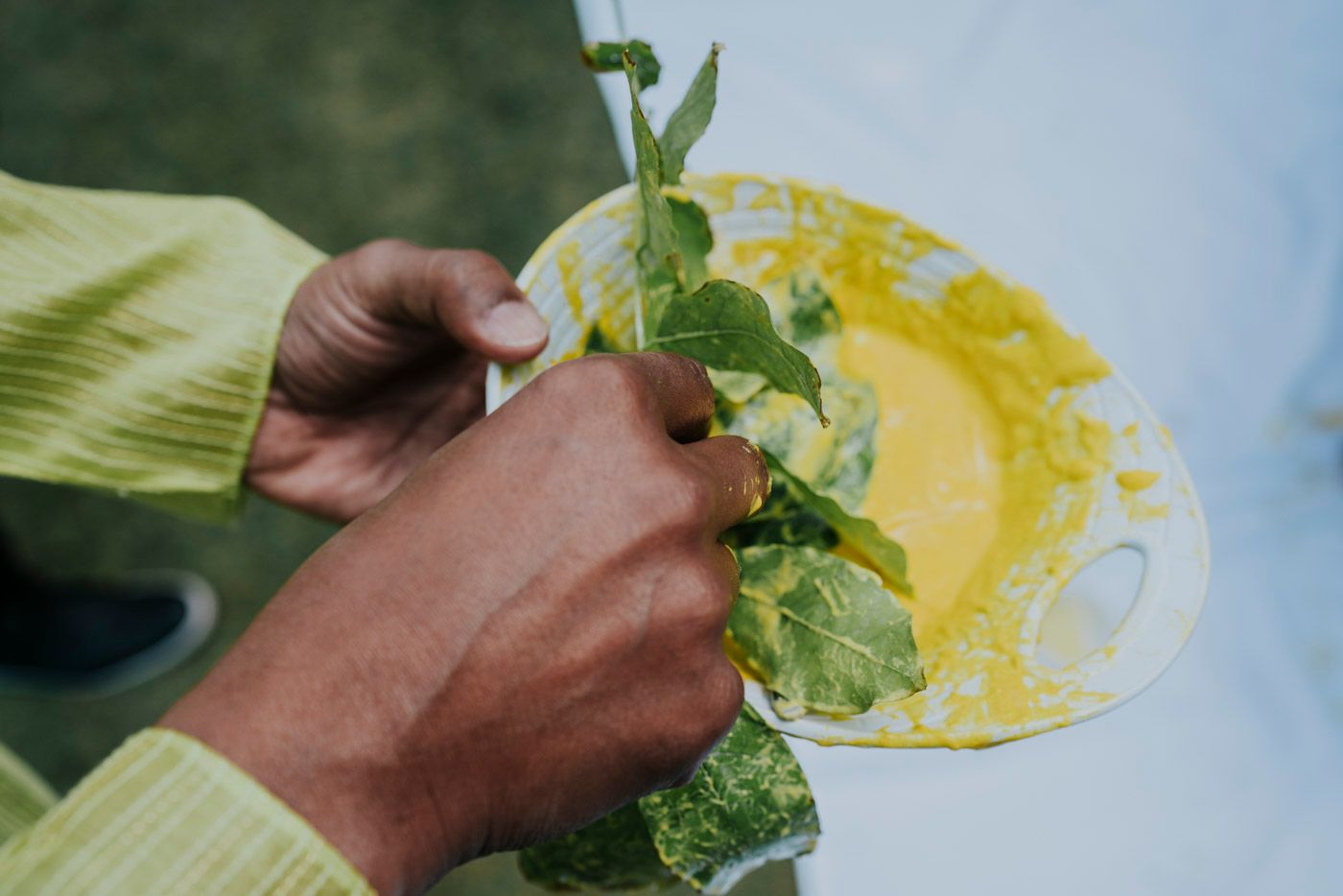 Hands gently prepare fresh salad ingredients over a yellow plate against a light blue background.