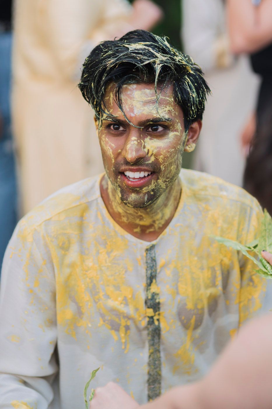 Person wearing a yellow stained shirt smiles while covered in green colored powder or paint during a festive celebration.