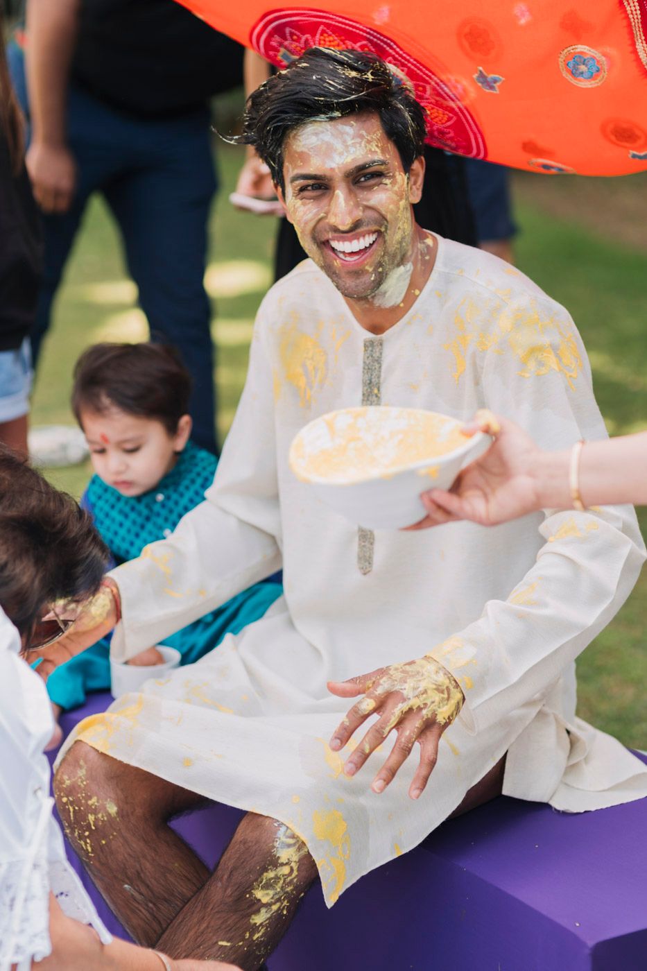 A person in a white kurta with yellow stains celebrates Holi festival while holding a beverage cup outdoors.