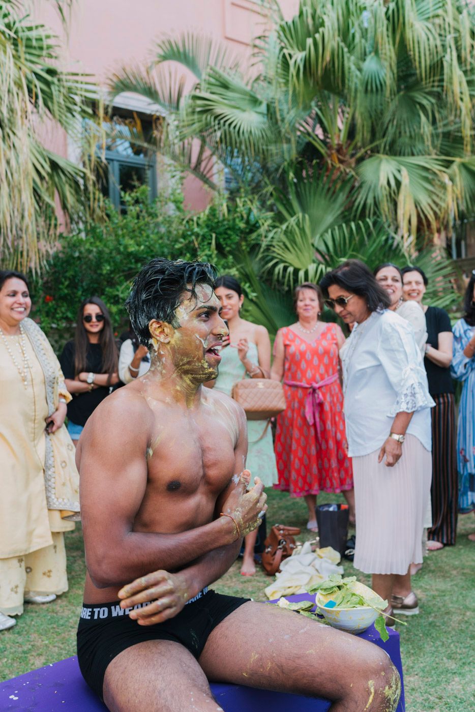 A traditional ceremonial gathering takes place in a lush garden setting with palm trees and tropical greenery.
