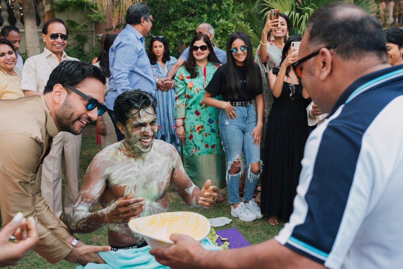 A festive outdoor gathering shows people celebrating with colorful powder in a garden setting.
