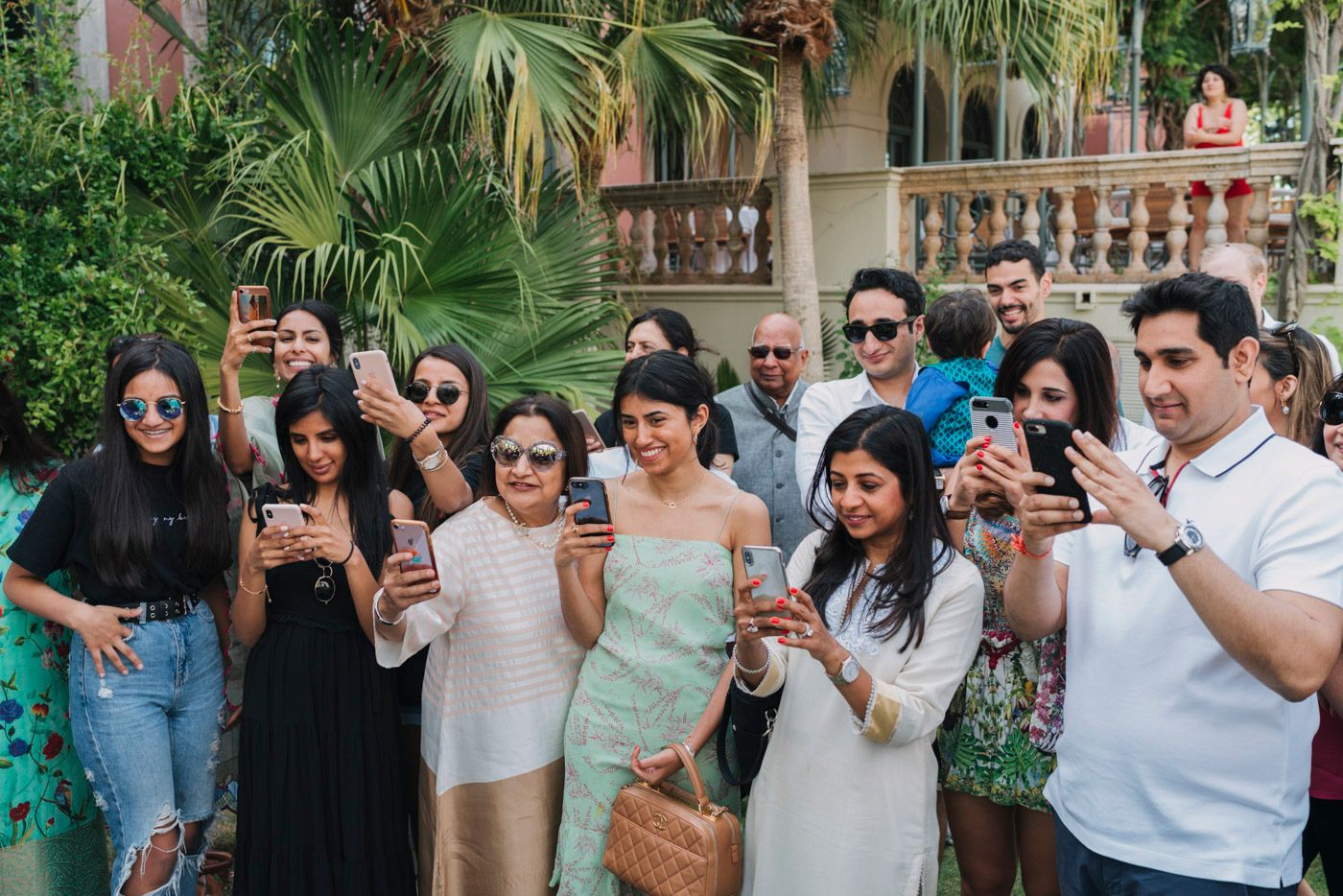 A group of friends standing together using their phones to take photos at an outdoor event with palm trees.