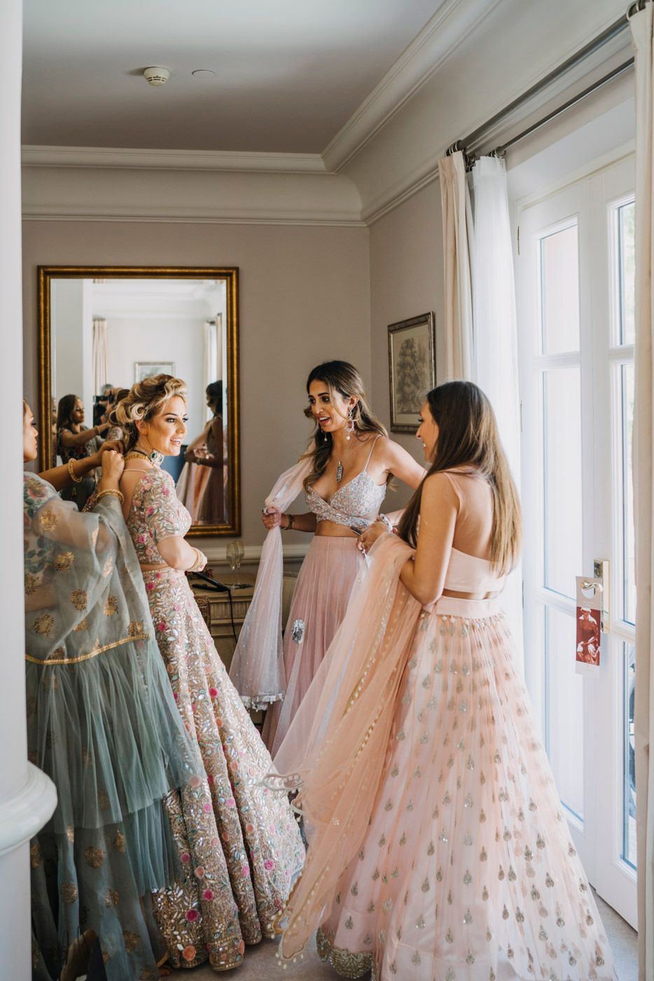 Three people in flowing formal gowns gather near a window in a bright room with elegant decor.