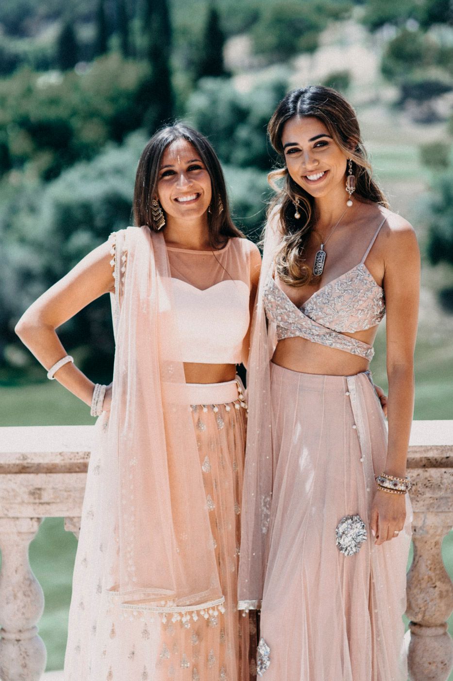 Two women pose together wearing modern blush pink Indian lehenga sets with crop tops and flowing skirts.