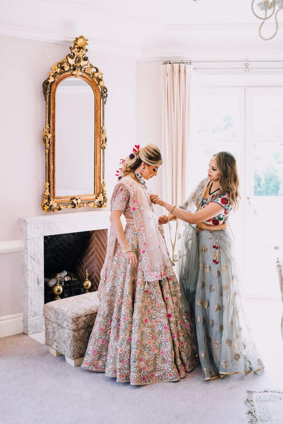 Two women in ornate floral gowns stand by an elegant fireplace with gold mirror in a bright white room.