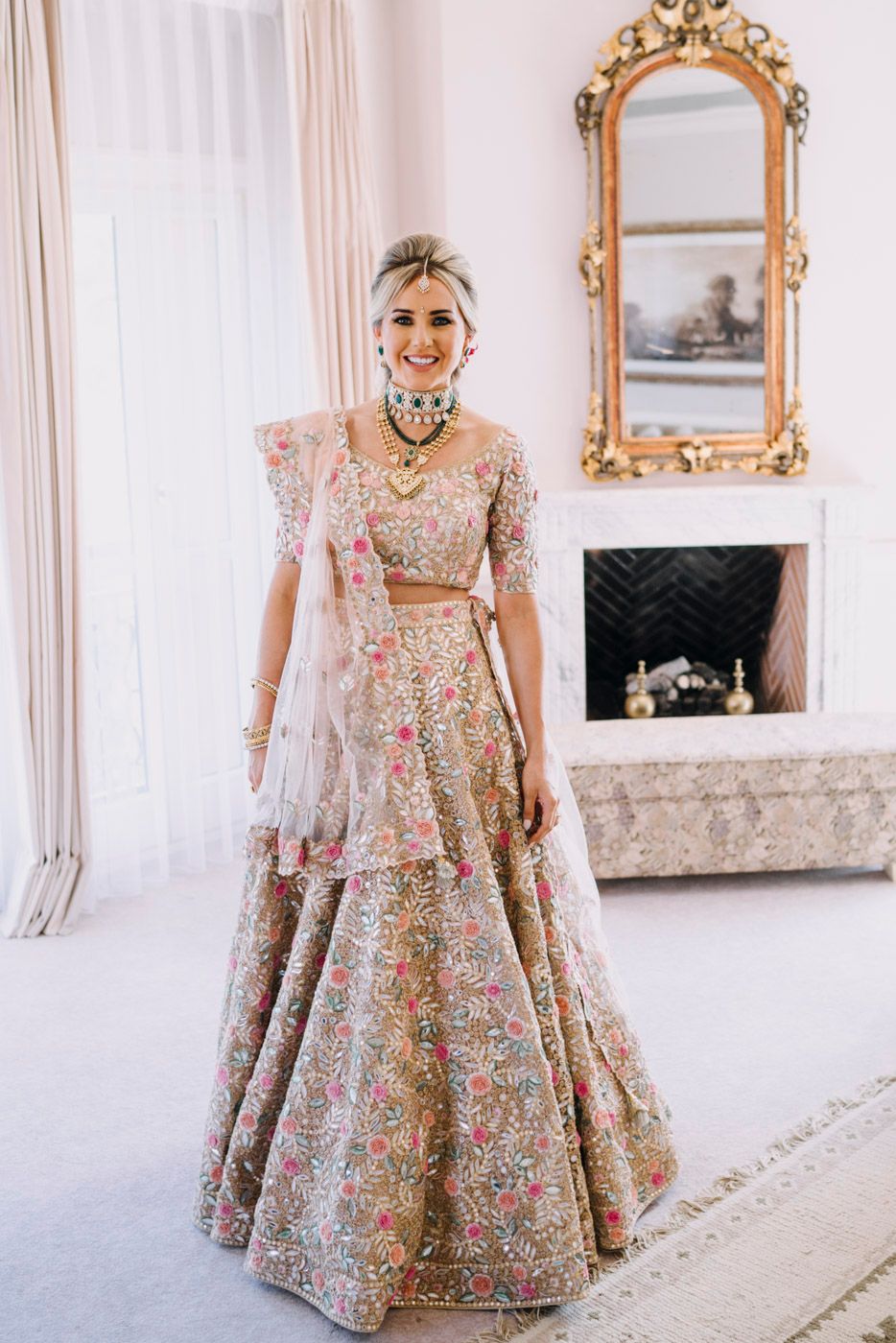 Elegant floral lehenga with intricate embroidery shown in luxurious setting with ornate gold mirror and fireplace.