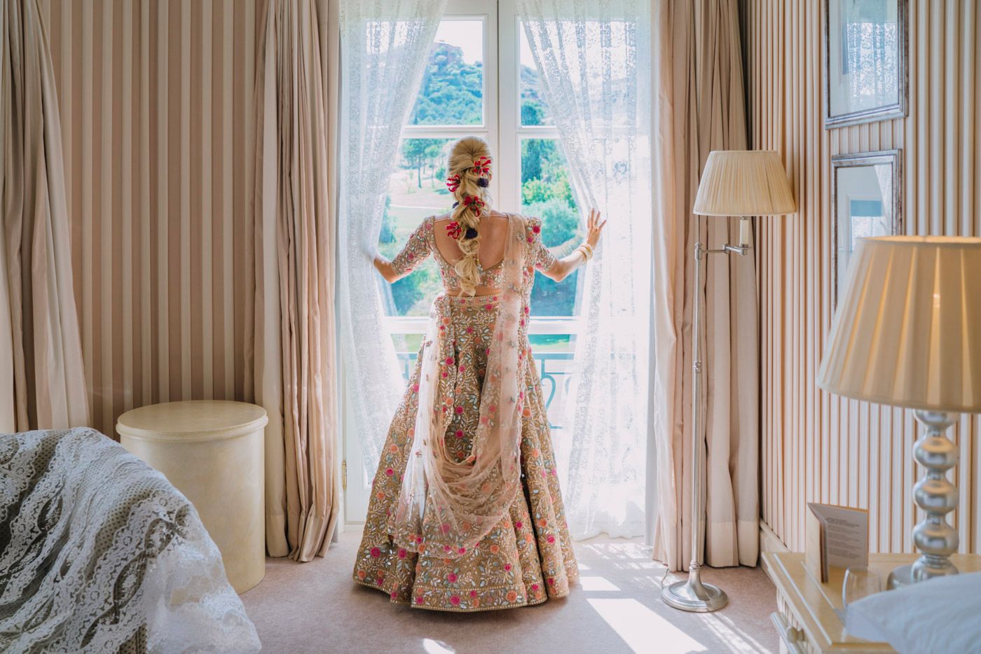 Elegant bridal attire featuring a detailed blush and gold lehenga with intricate embroidery by a window with sheer curtains.