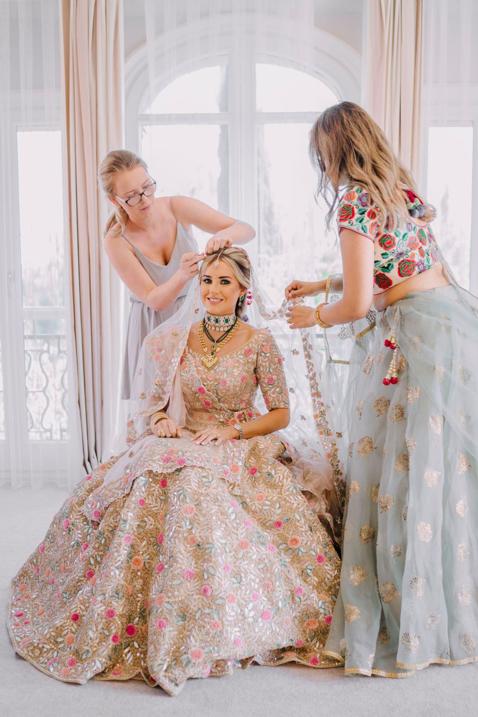 Final preparations of a bride wearing an embellished pink lehenga in an elegant room with arched windows.