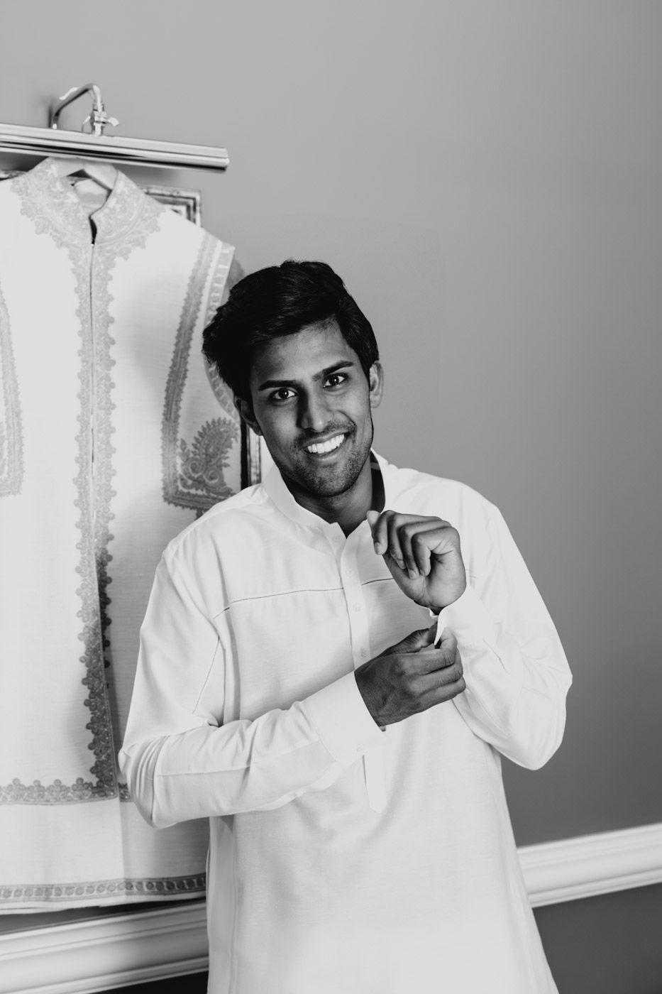 Smiling person wearing white traditional kurta adjusts button while standing against a gray background.