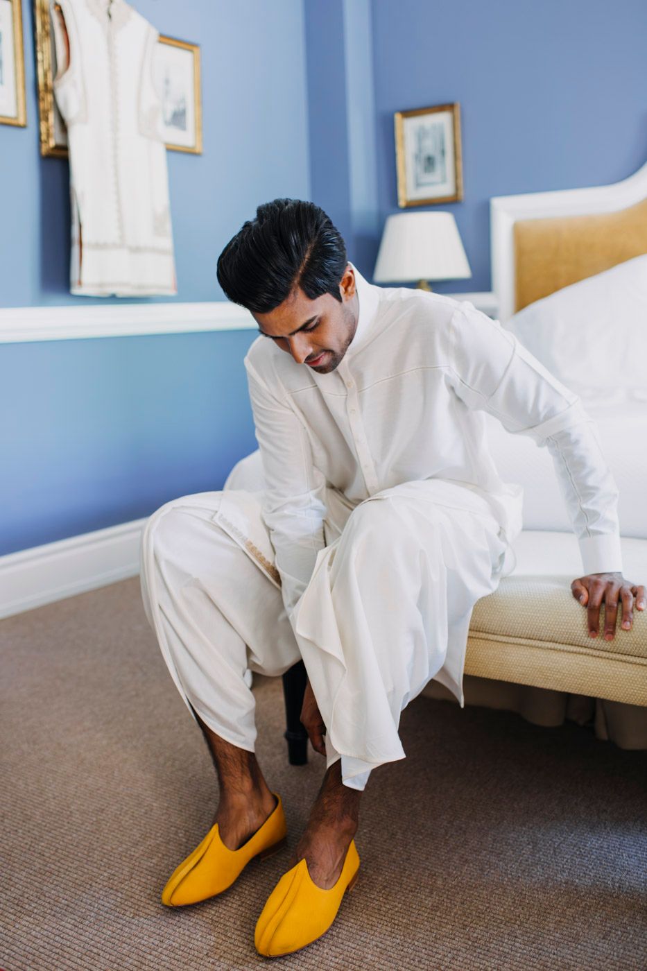 Someone in all-white attire sits on a bed wearing bright yellow slippers in a room with blue walls.