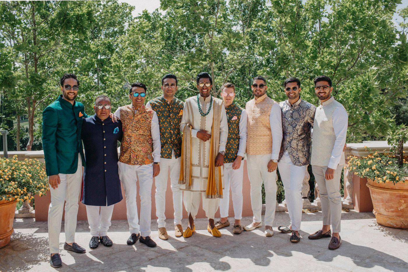 A group stands together in formal ethnic attire against a garden backdrop with white pants and varied colored tops.