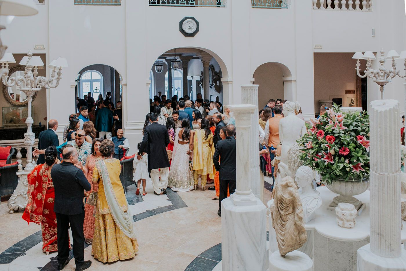Guests mingle in an elegant white ballroom with ornate columns and chandeliers during an upscale event.