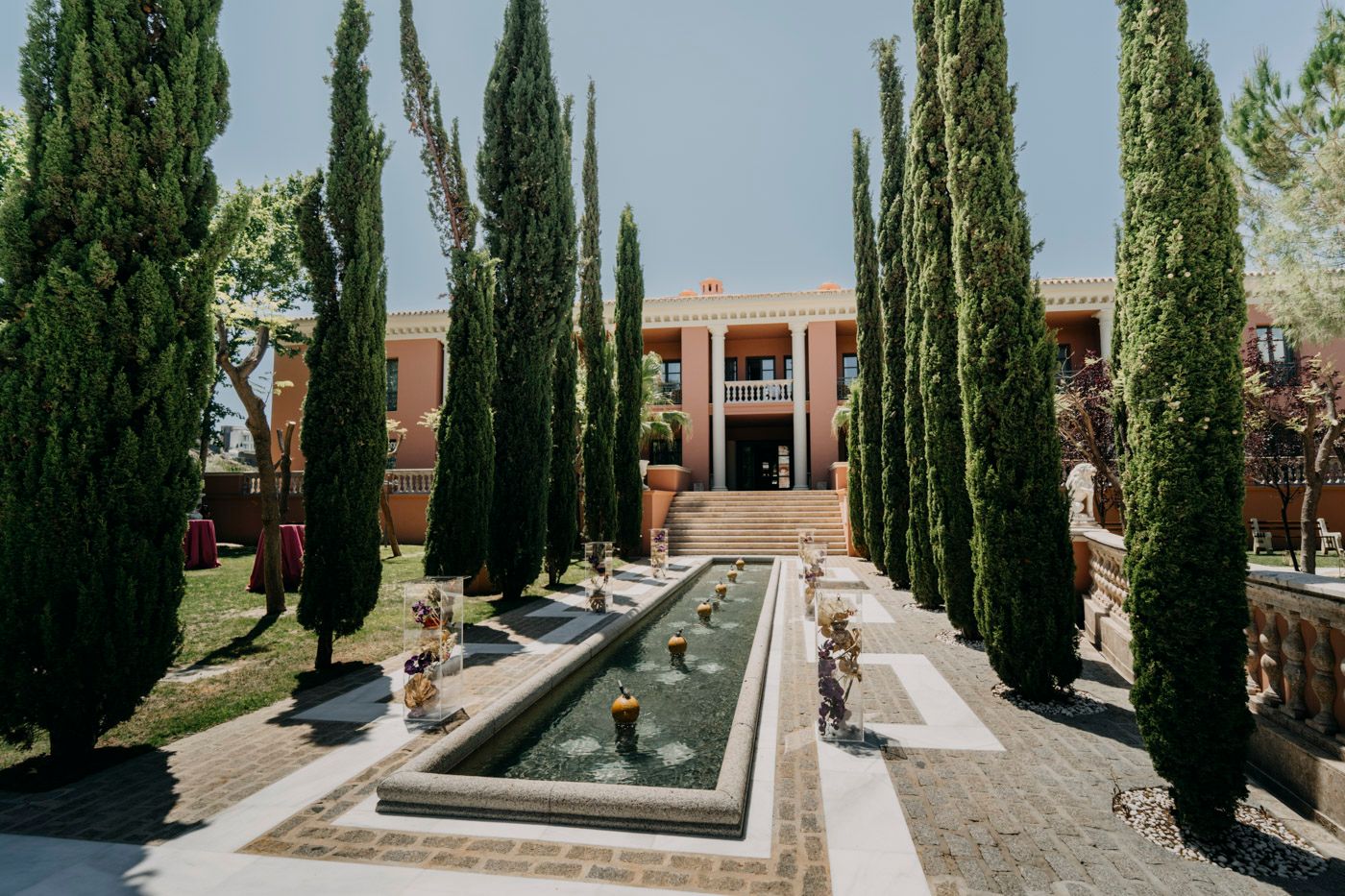 Mediterranean style courtyard with tall cypress trees flanking a rectangular reflecting pool and brick building.