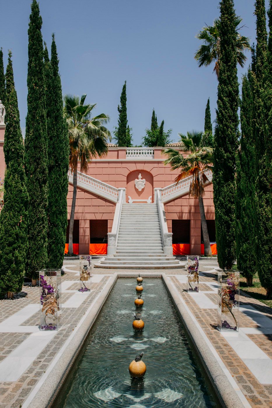 Mediterranean-style courtyard features grand white stairs, cypress trees, and reflecting pool with floating golden spheres.