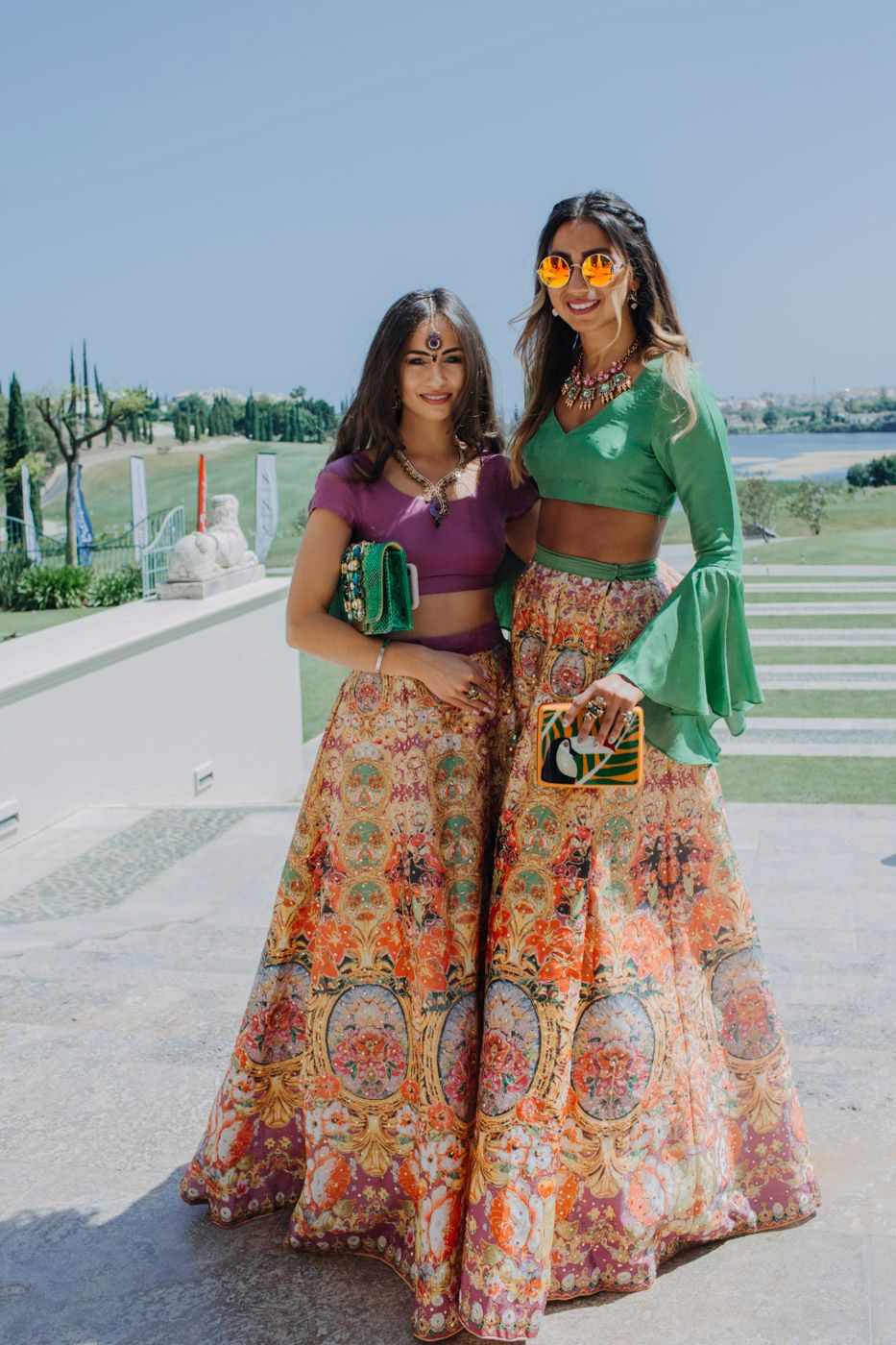 Two people in colorful Indian lehengas with floral patterns pose outdoors wearing crop tops and matching skirts.
