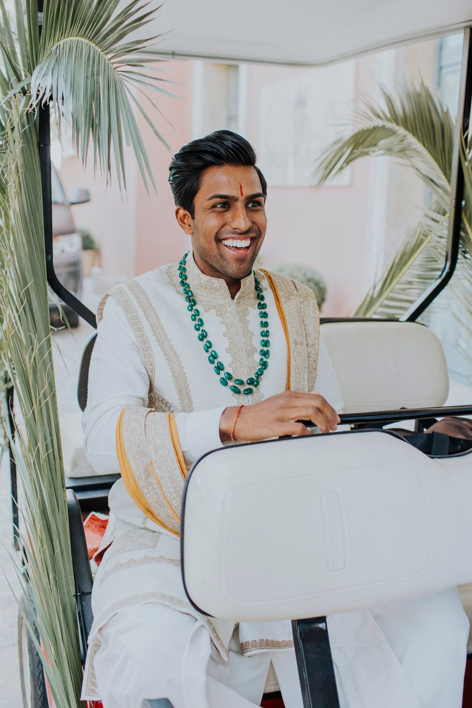 A person in a white traditional outfit with green beaded necklace sits smiling while driving a golf cart among palm trees.