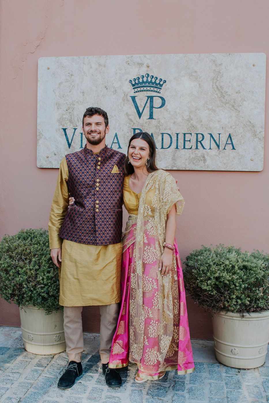Two people in traditional Indian attire with maroon and yellow colors pose in front of a Villa Pasterna sign.