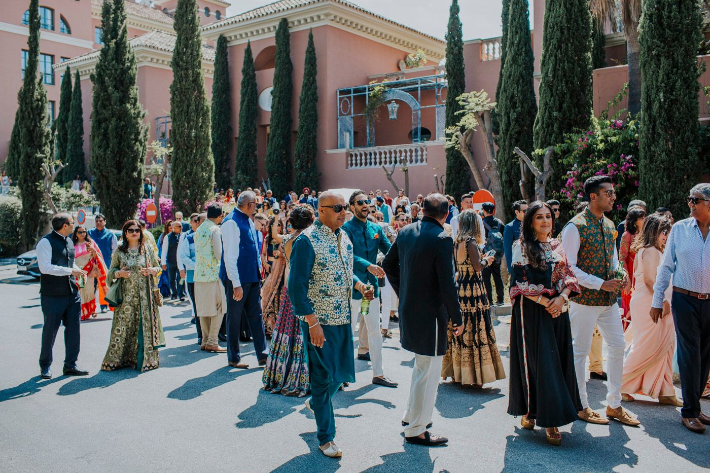 A crowd of people gather outdoors in a Mediterranean setting with cypress trees and pink buildings on a sunny day.