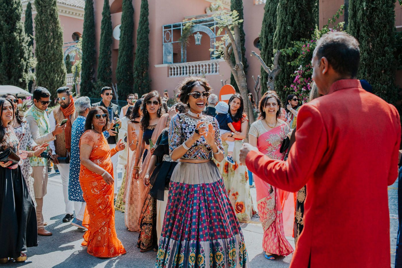 Guests in colorful traditional Indian wedding attire dance and celebrate at an outdoor venue with cypress trees in background.