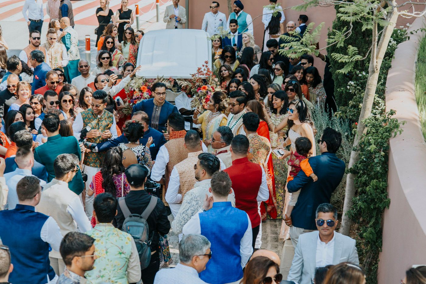 Large crowd of people gathered at an outdoor event with a sign visible in the background against pink walls.