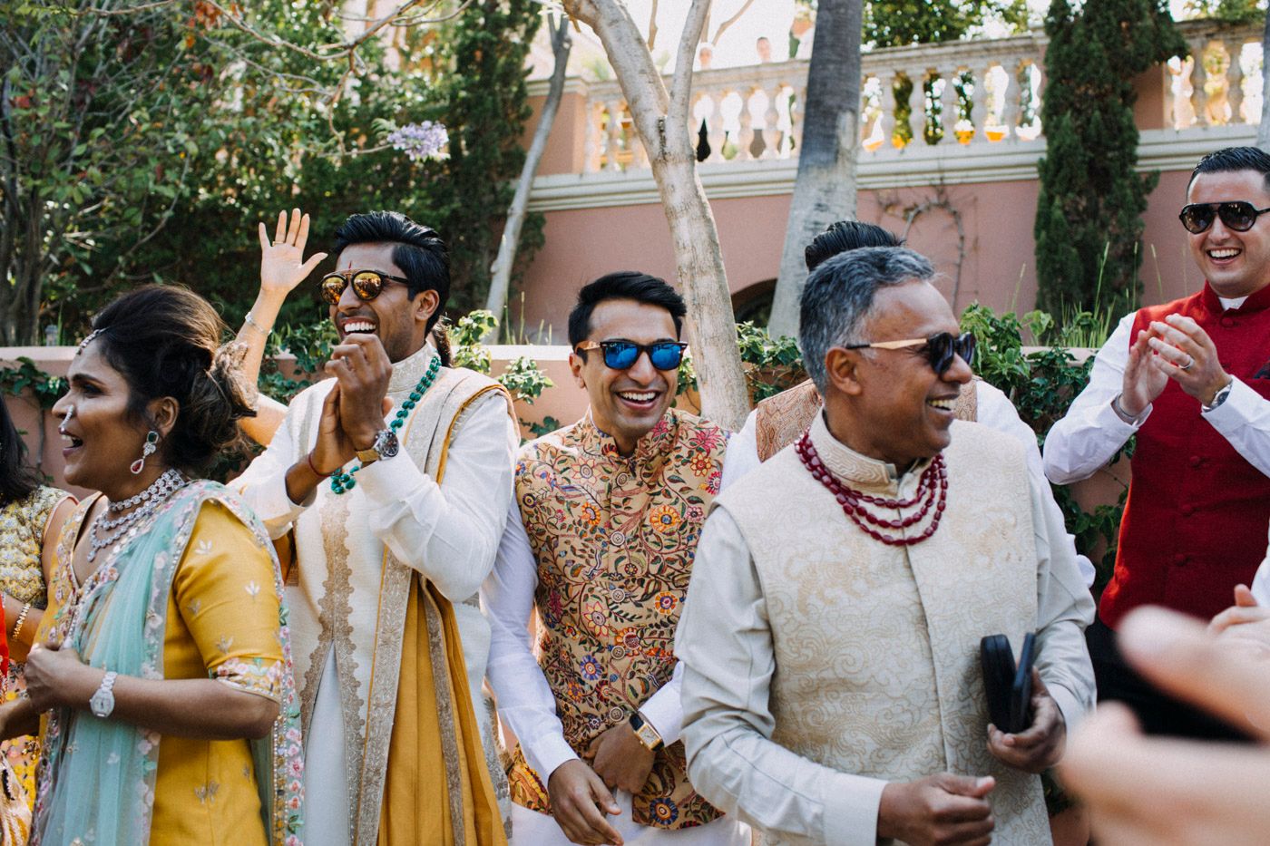 A group of festively dressed people dancing and celebrating at an outdoor event with trees and buildings in the background.