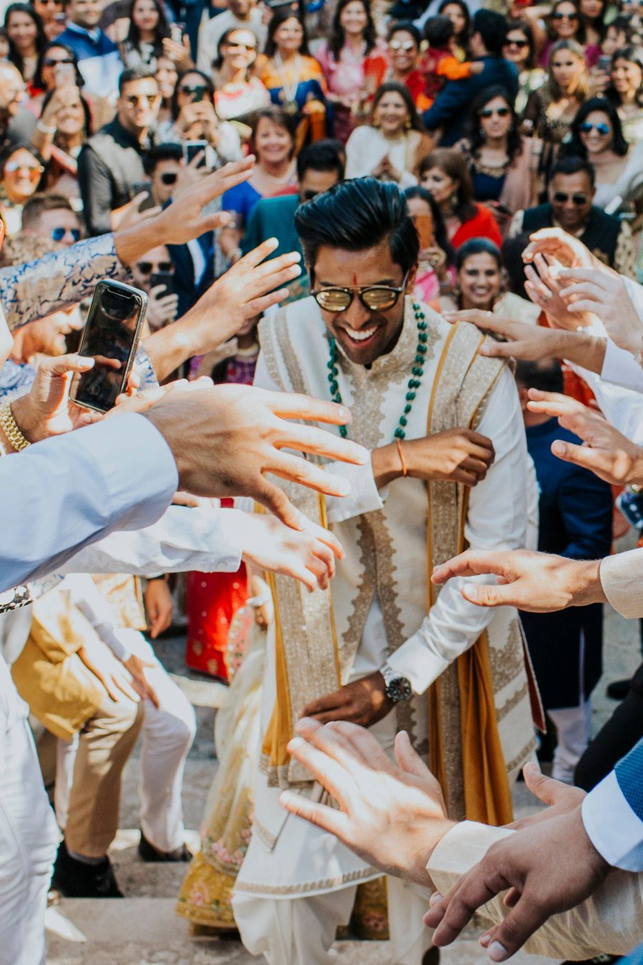 A joyous participant in traditional Indian wedding attire is surrounded by reaching hands during a celebratory moment.