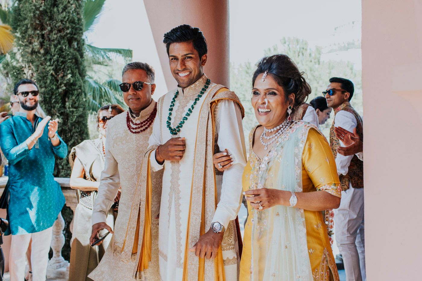 Guests wearing traditional Indian attire in yellow, white and turquoise celebrate at a cultural wedding ceremony outdoors.