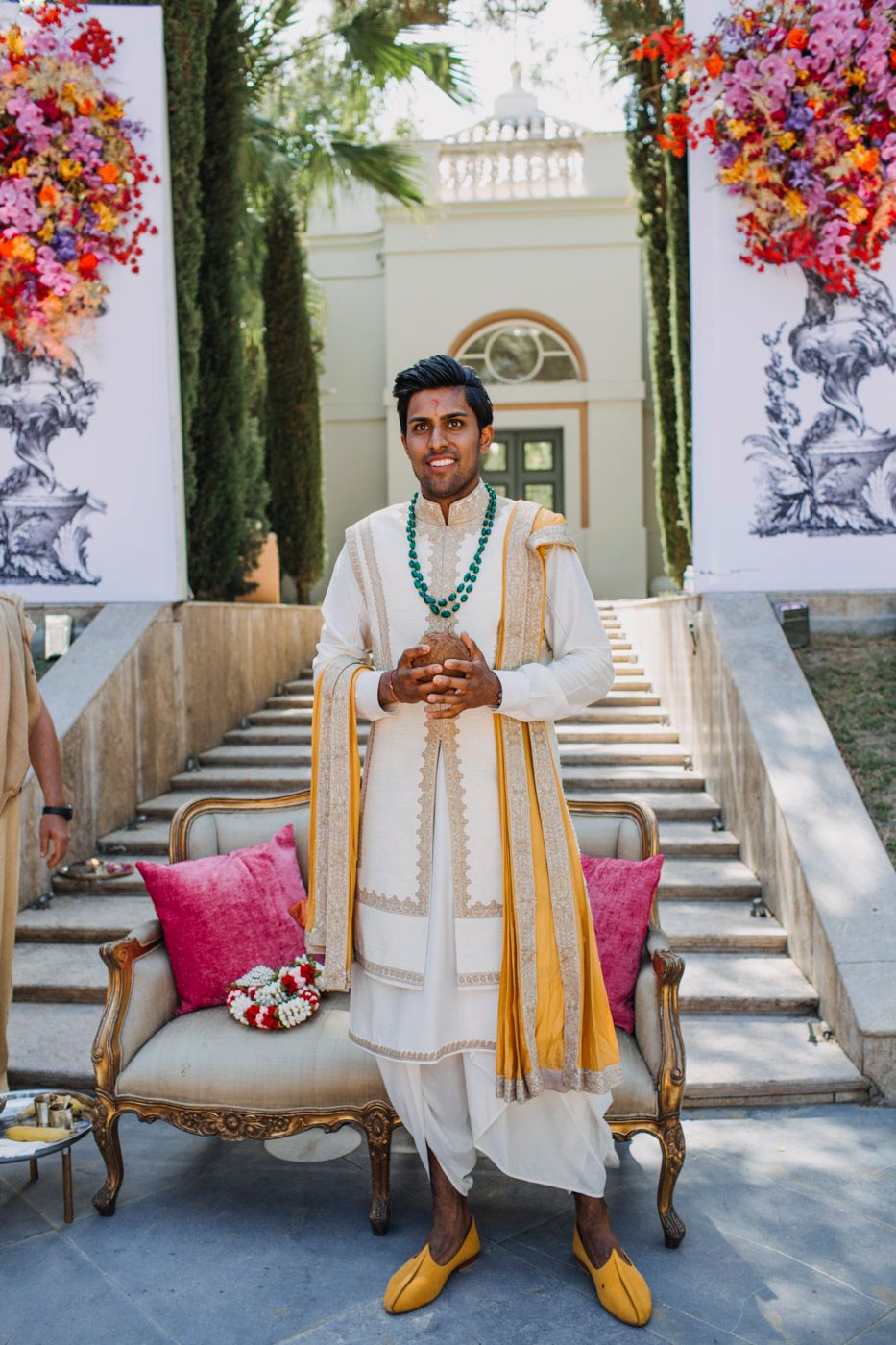 Traditional Indian groom stands at elegant staircase with pink floral decor wearing cream sherwani and turquoise necklace.