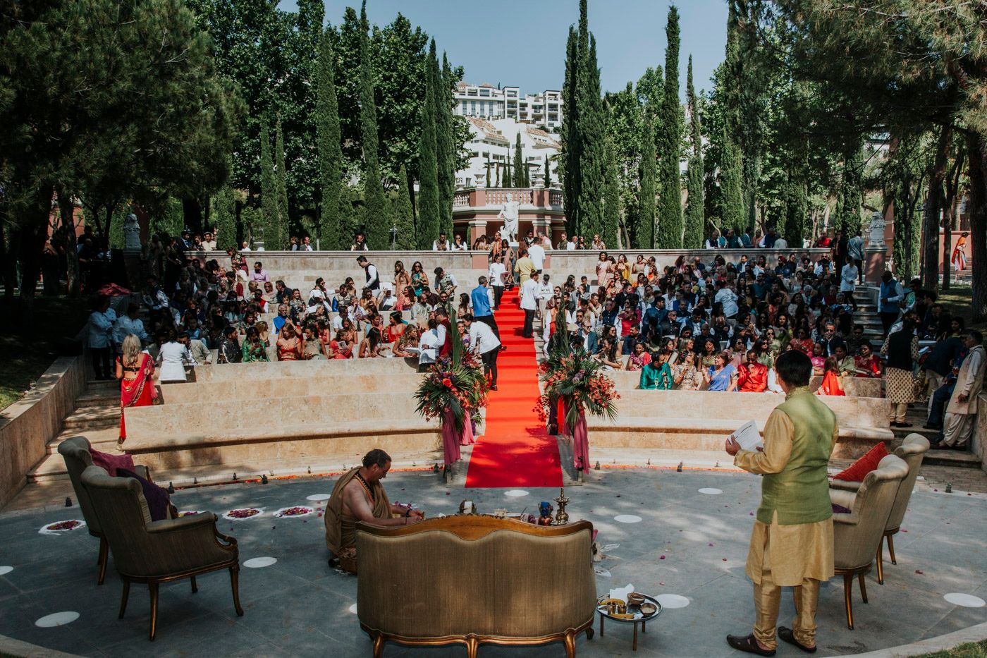 An outdoor performance event with two pianos and a red carpet in a garden setting surrounded by tall cypress trees.