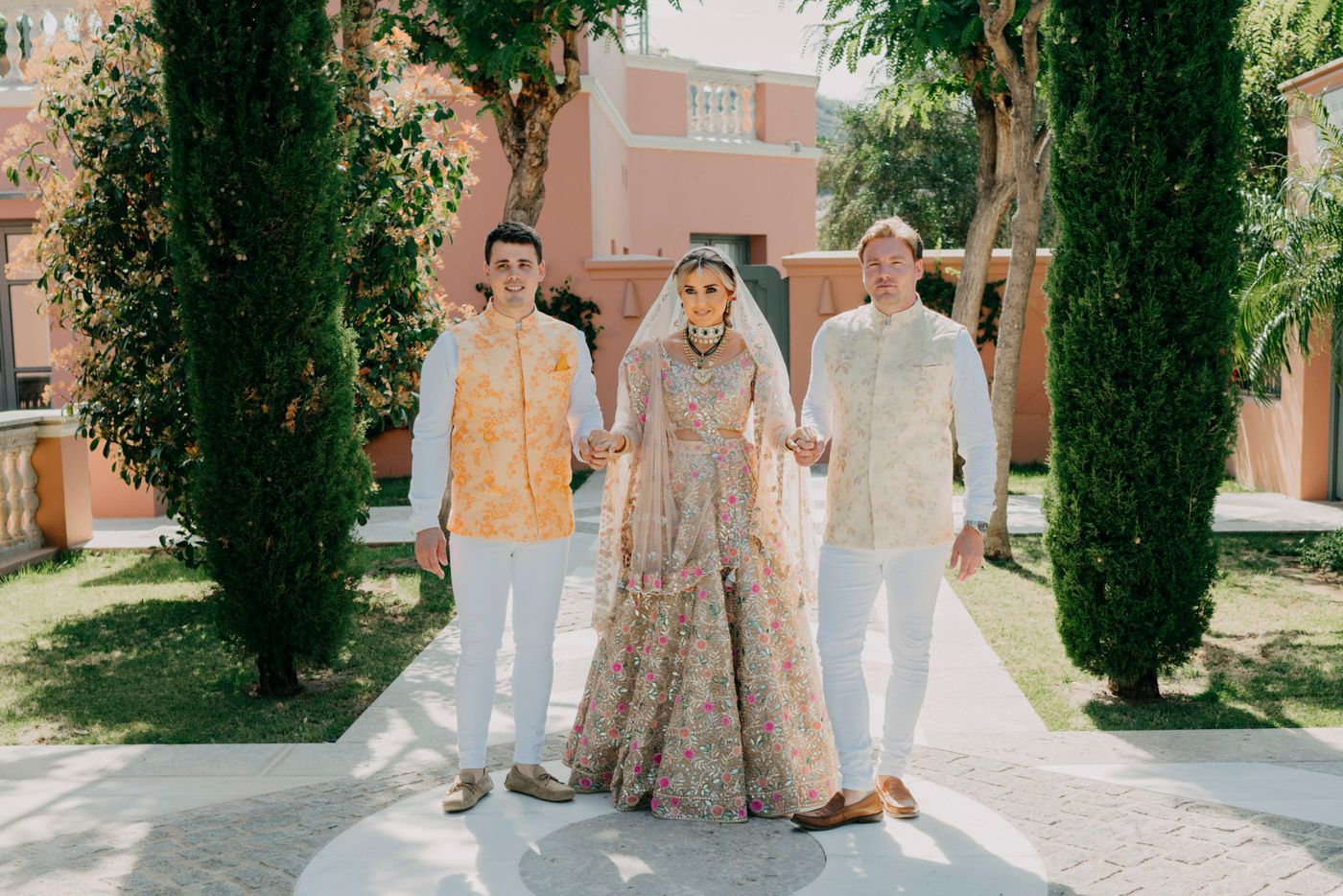 Three people in formal attire standing on a path between cypress trees outside a pink building.