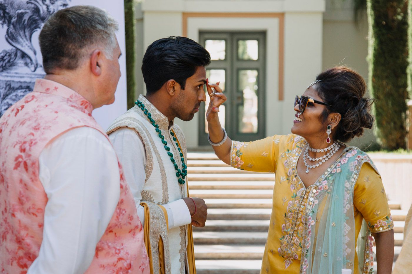 Traditional Indian wedding ceremony with three people in colorful formal attire interacting on outdoor steps.