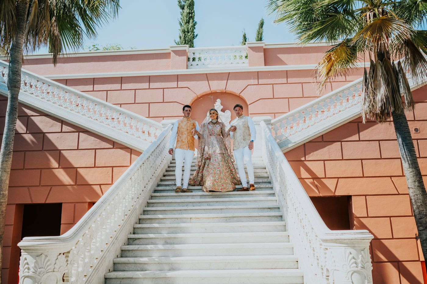 Elegant white staircase with ornate railings leads up to a coral-colored building with palm trees in the background.