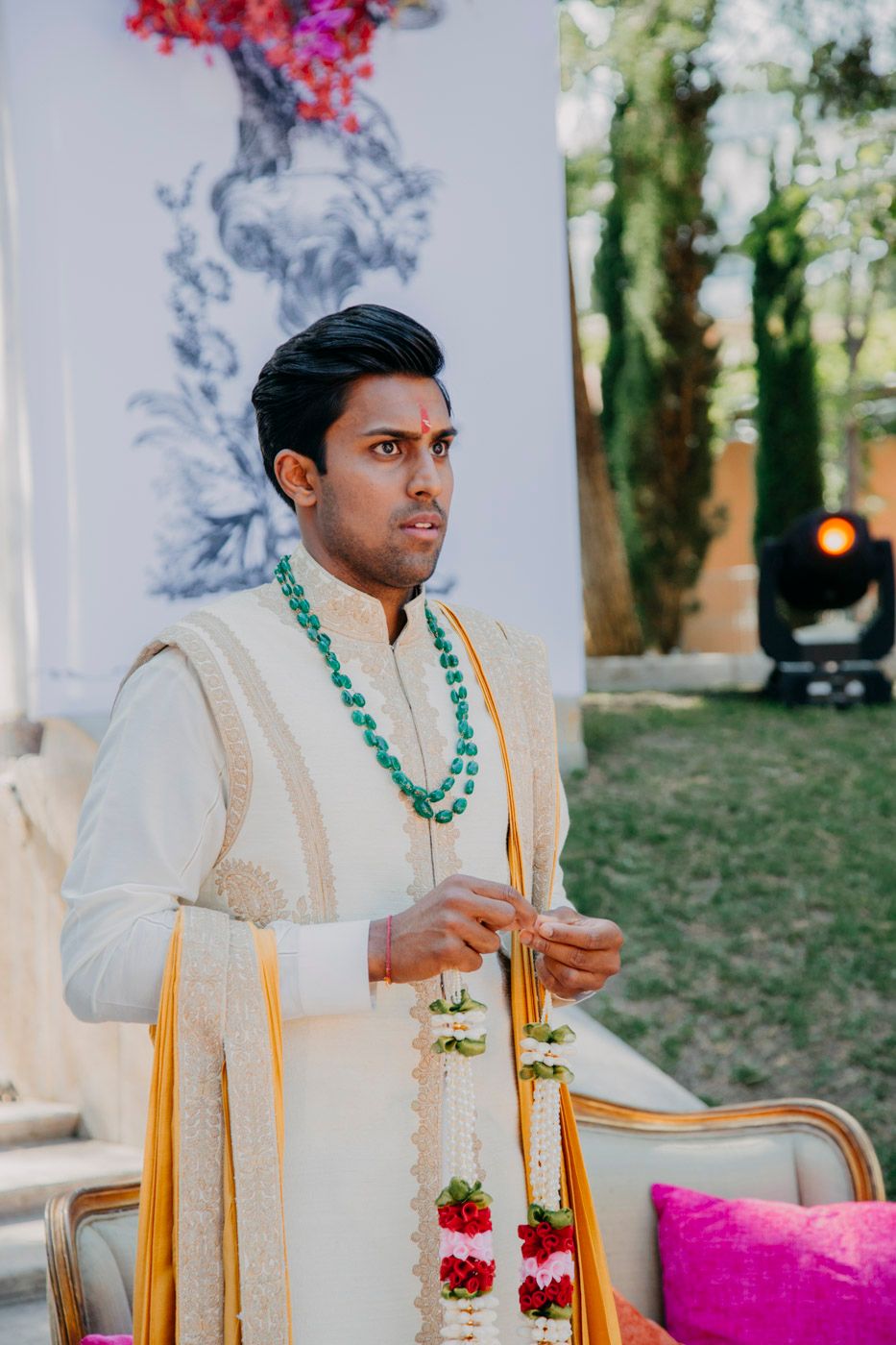 Traditional Indian groom wearing cream sherwani and green beaded necklace during wedding ceremony.