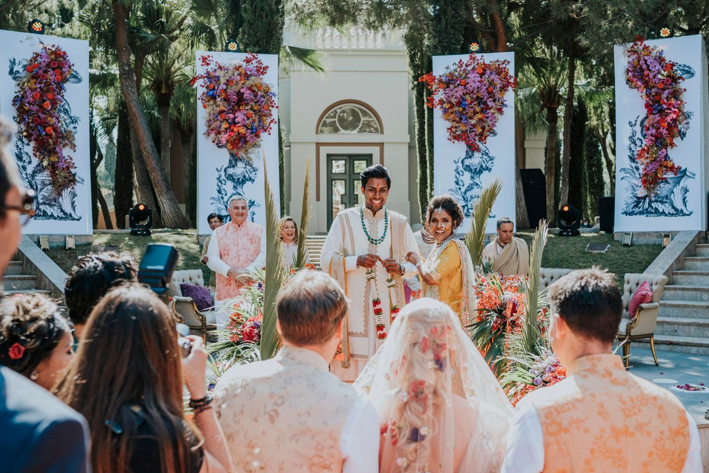 An outdoor wedding ceremony decorated with vibrant red floral banners and lush greenery in a garden setting.