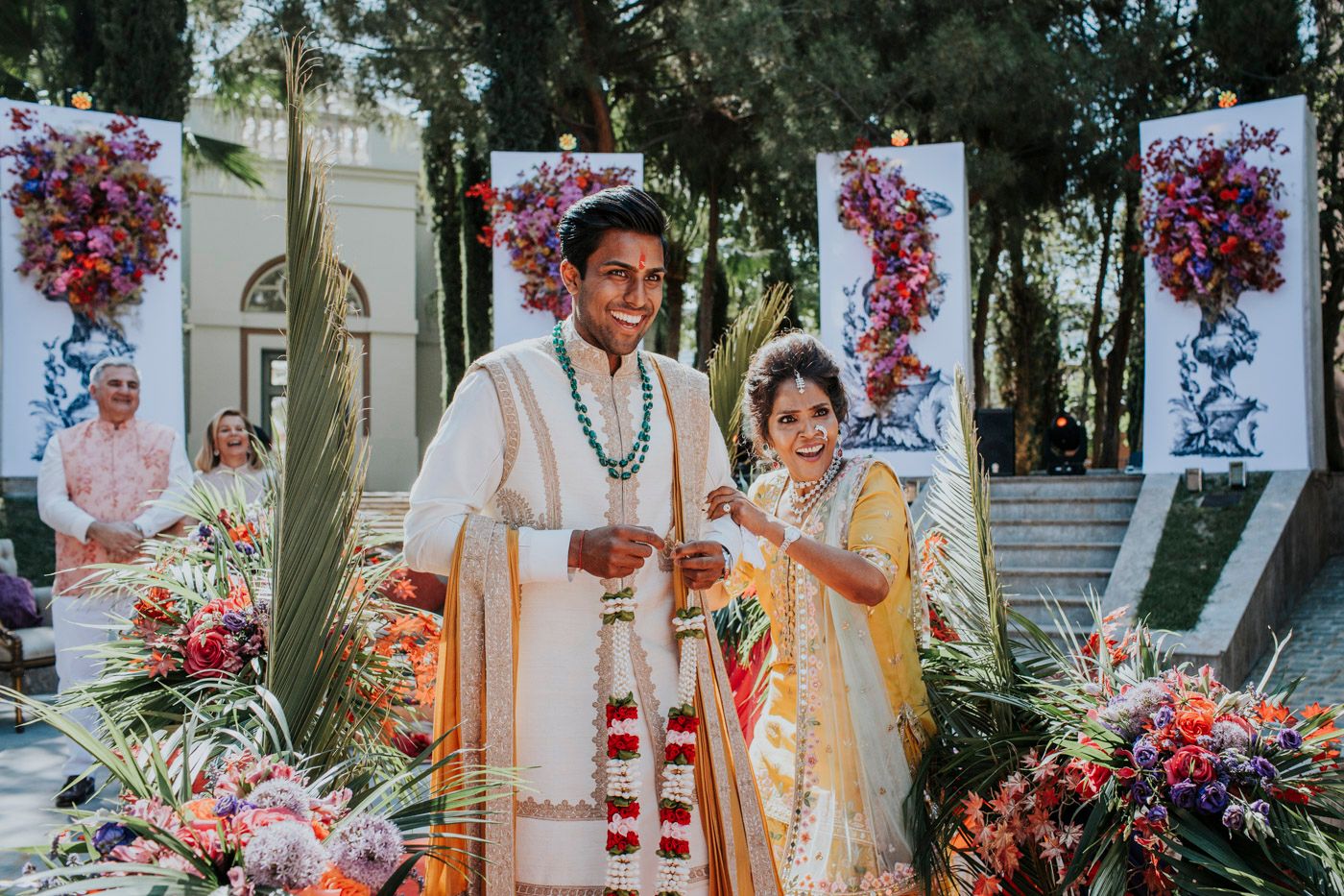 A traditional outdoor Indian wedding ceremony with festive floral decorations and pink flowers hanging in the background.