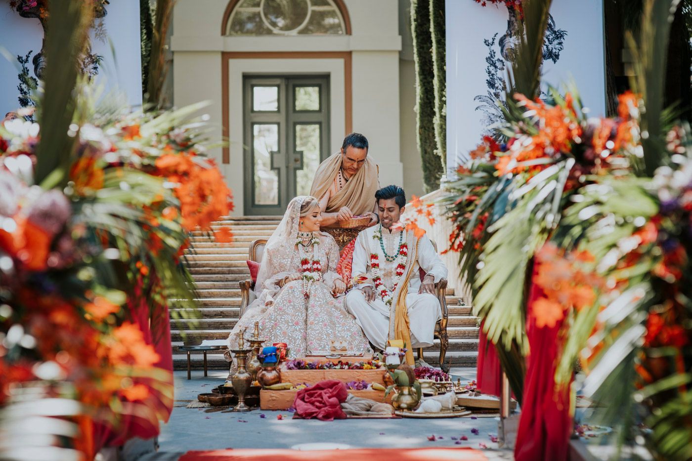 Traditional Indian wedding ceremony on outdoor steps surrounded by vibrant orange and red floral decorations.