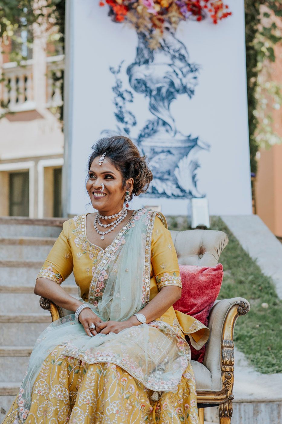 Traditional South Asian bride in yellow and gold lehenga poses elegantly on ornate chair during wedding photoshoot.