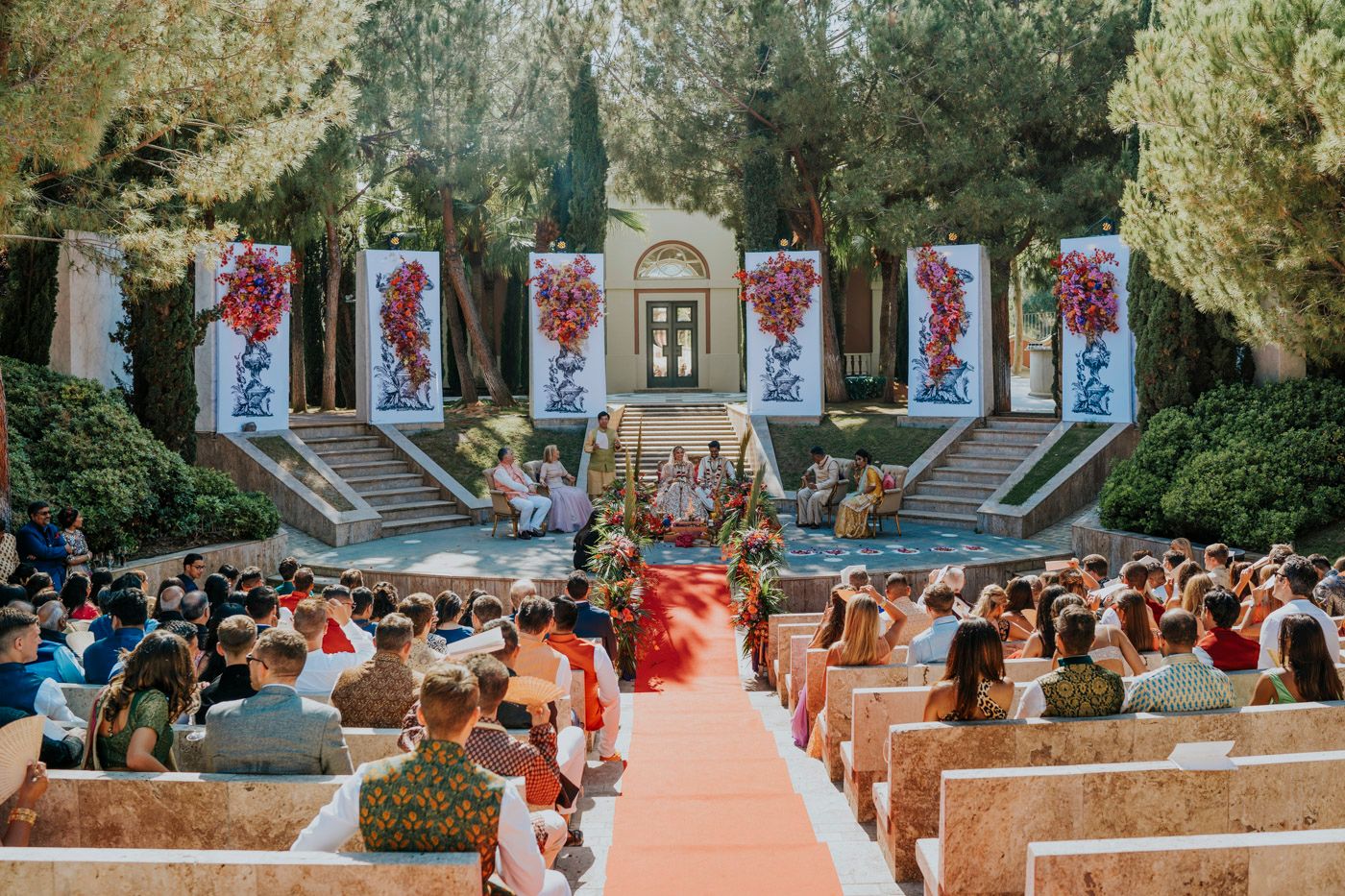 An outdoor wedding ceremony at an amphitheater with red and white banners decorating stone columns along the aisle.