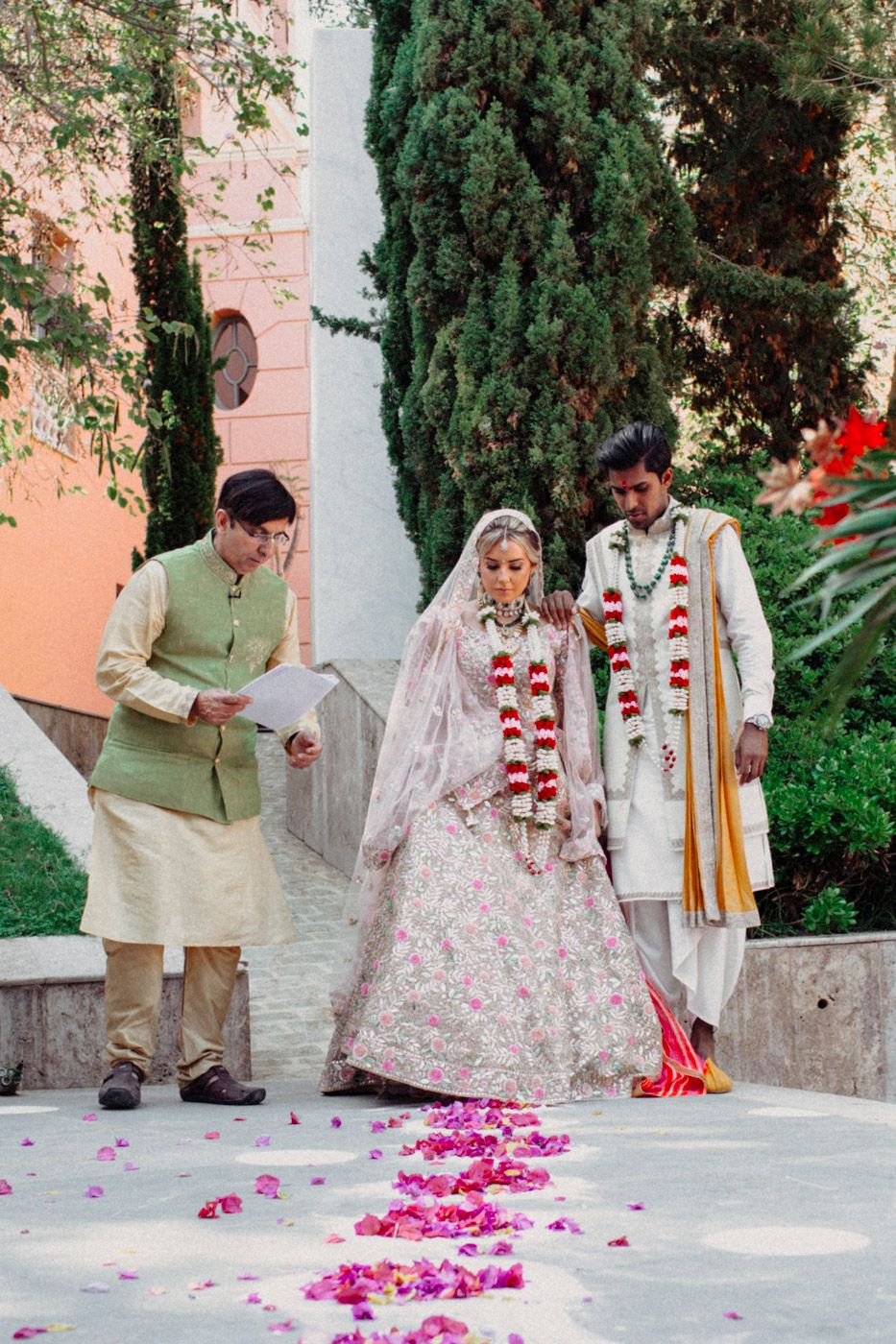 Wedding ceremony in garden setting with traditional South Asian bridal couple wearing garlands and officiant in green.