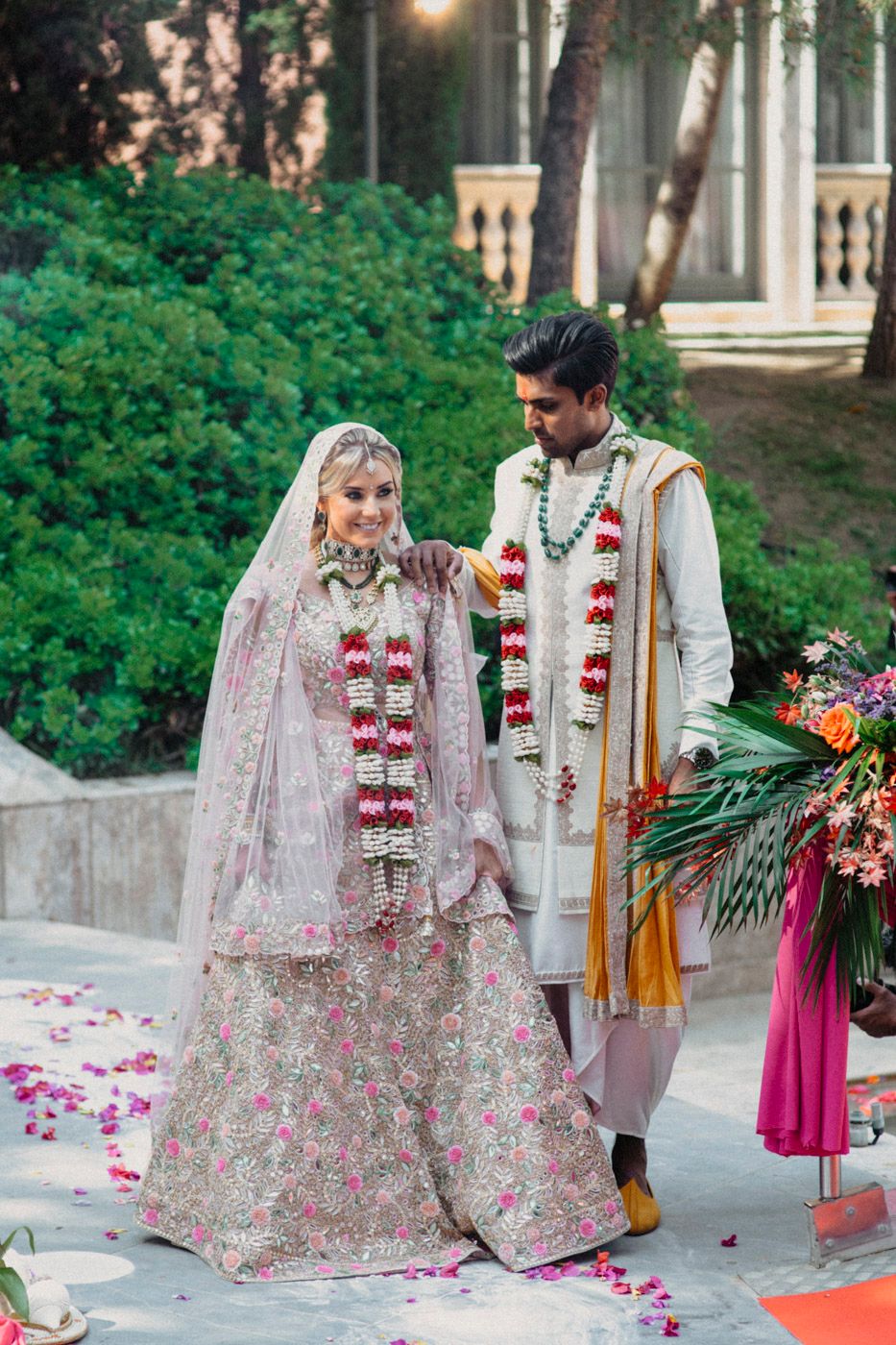 A couple in traditional Indian wedding attire stands among lush greenery wearing red and white floral garlands.