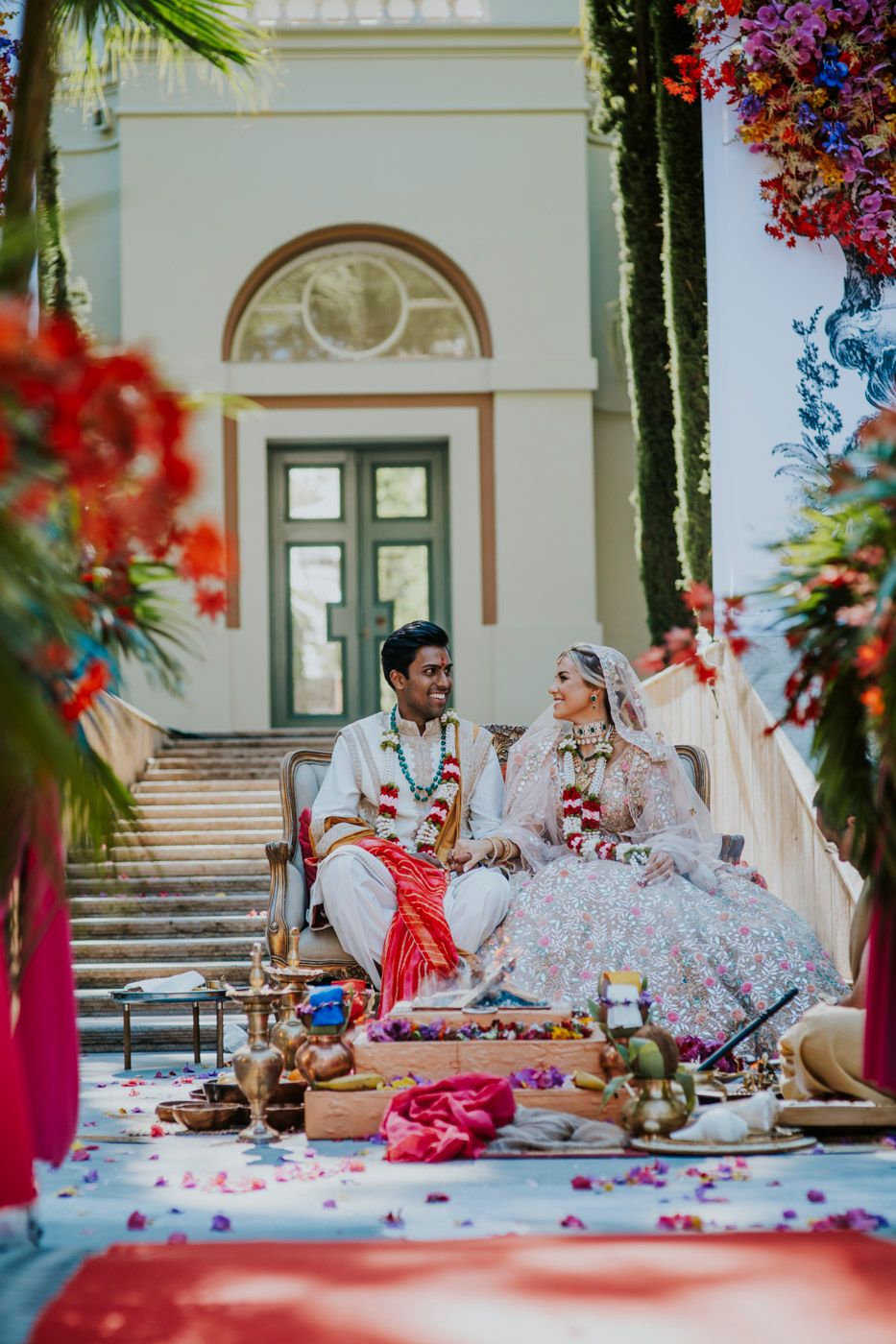 A couple in traditional South Asian wedding attire enjoy a decorated outdoor celebration with rose petals and ornate decor.
