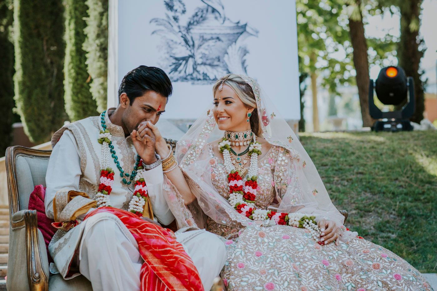 Wedding couple in traditional Indian attire share a candid moment while wearing floral garlands during outdoor ceremony.