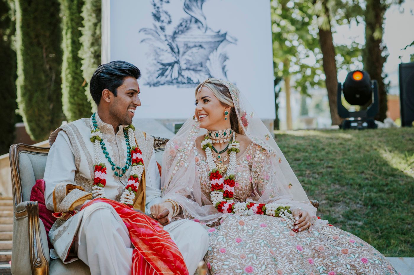 A couple in traditional wedding attire sits together outdoors surrounded by floral decorations and greenery.