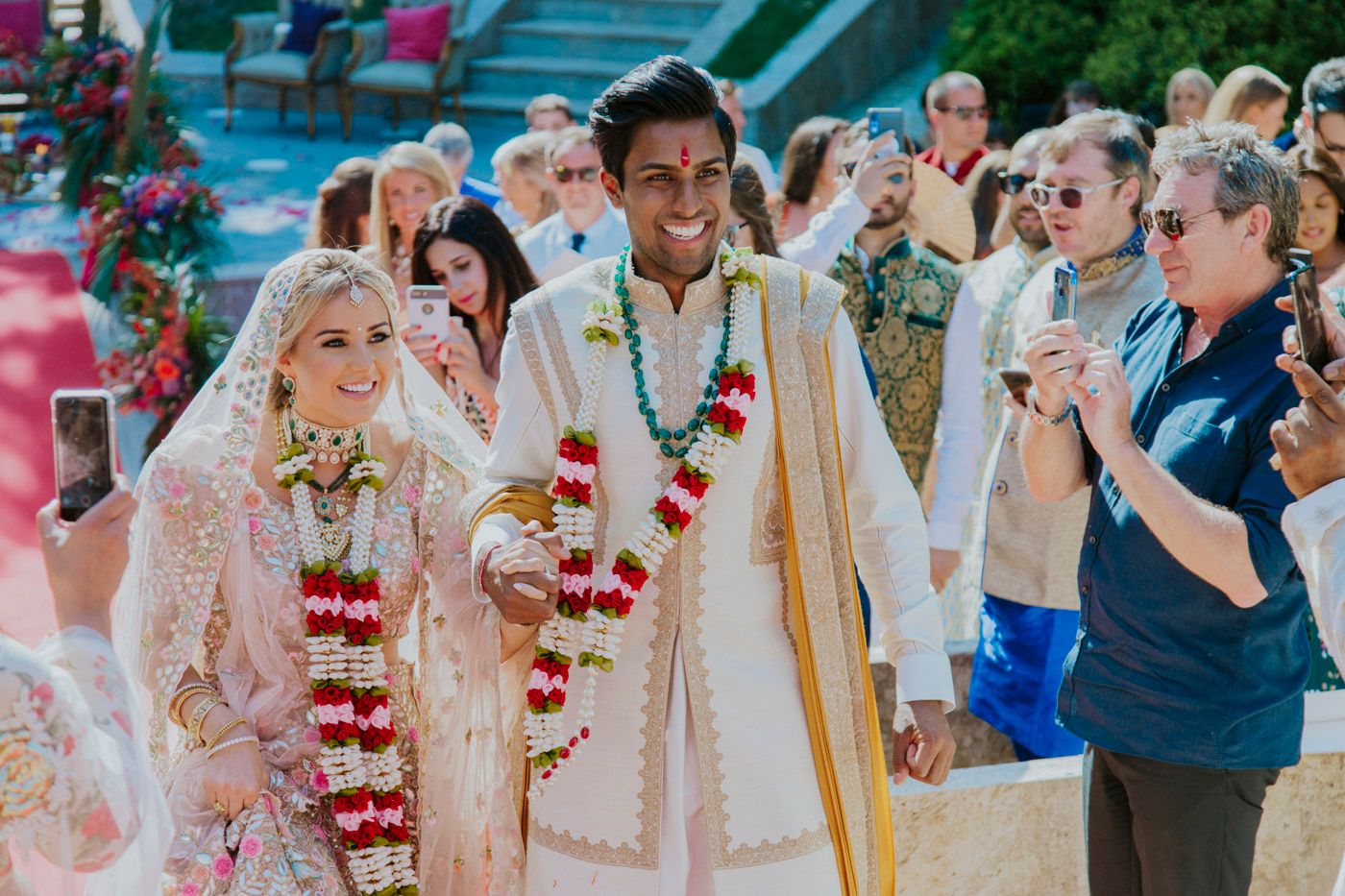 Wedding ceremony with couple in traditional white and gold Indian attire surrounded by guests outdoors.