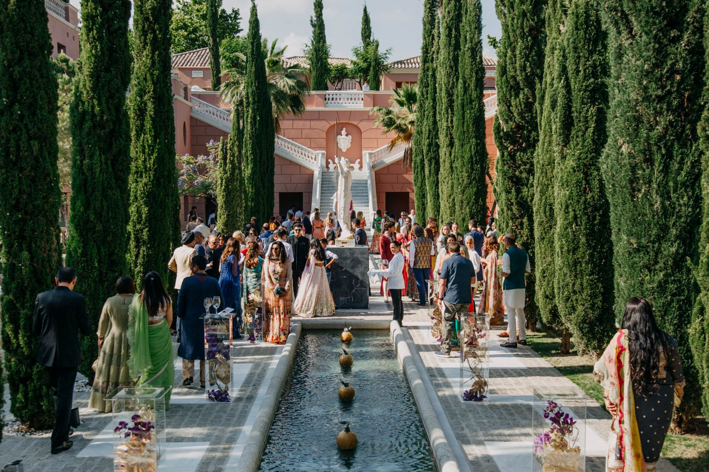 Formal garden ceremony aisle lined with tall cypress trees and reflecting pool at elegant destination wedding venue.