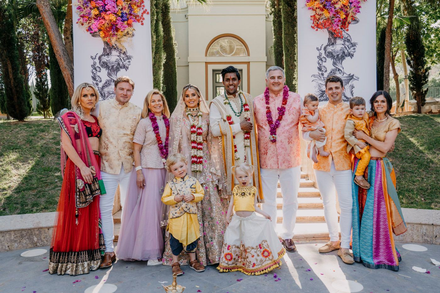 A festive wedding party wearing traditional Indian and fusion attire poses outdoors in front of a white building entrance.