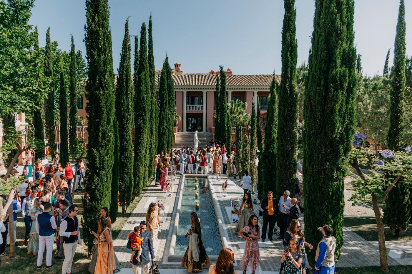 Crowds fill a pathway lined with tall cypress trees leading to an elegant mansion with architectural details.