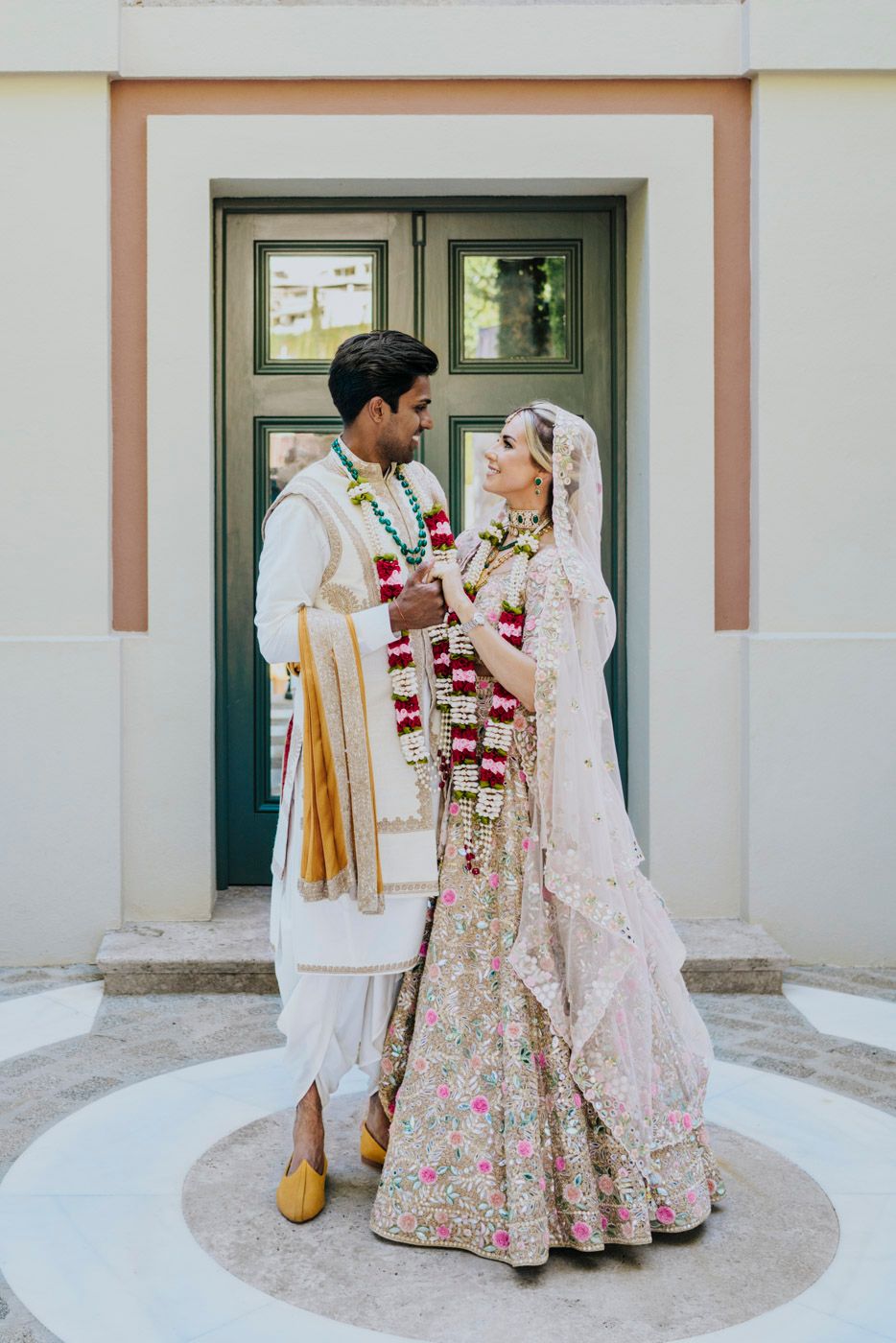 A couple in traditional Indian wedding attire stands together in front of a green doorway, with the bride in a floral lehenga.