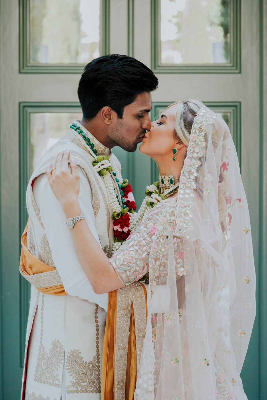 Romantic kiss between newlyweds wearing traditional Indian wedding attire against mint green doors.
